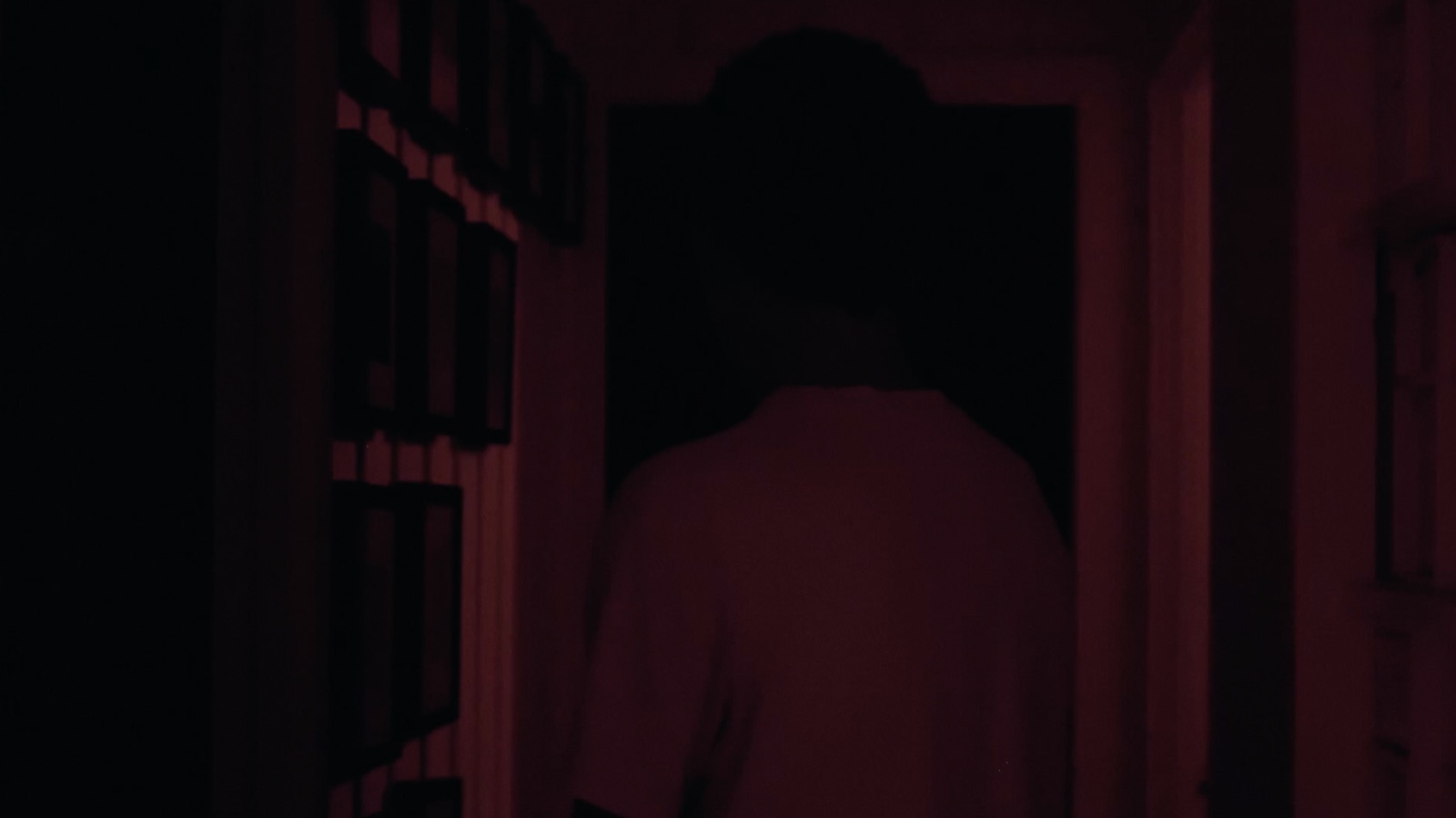 A figure walks away down a hallway lit deep red, framed photographs on the wall