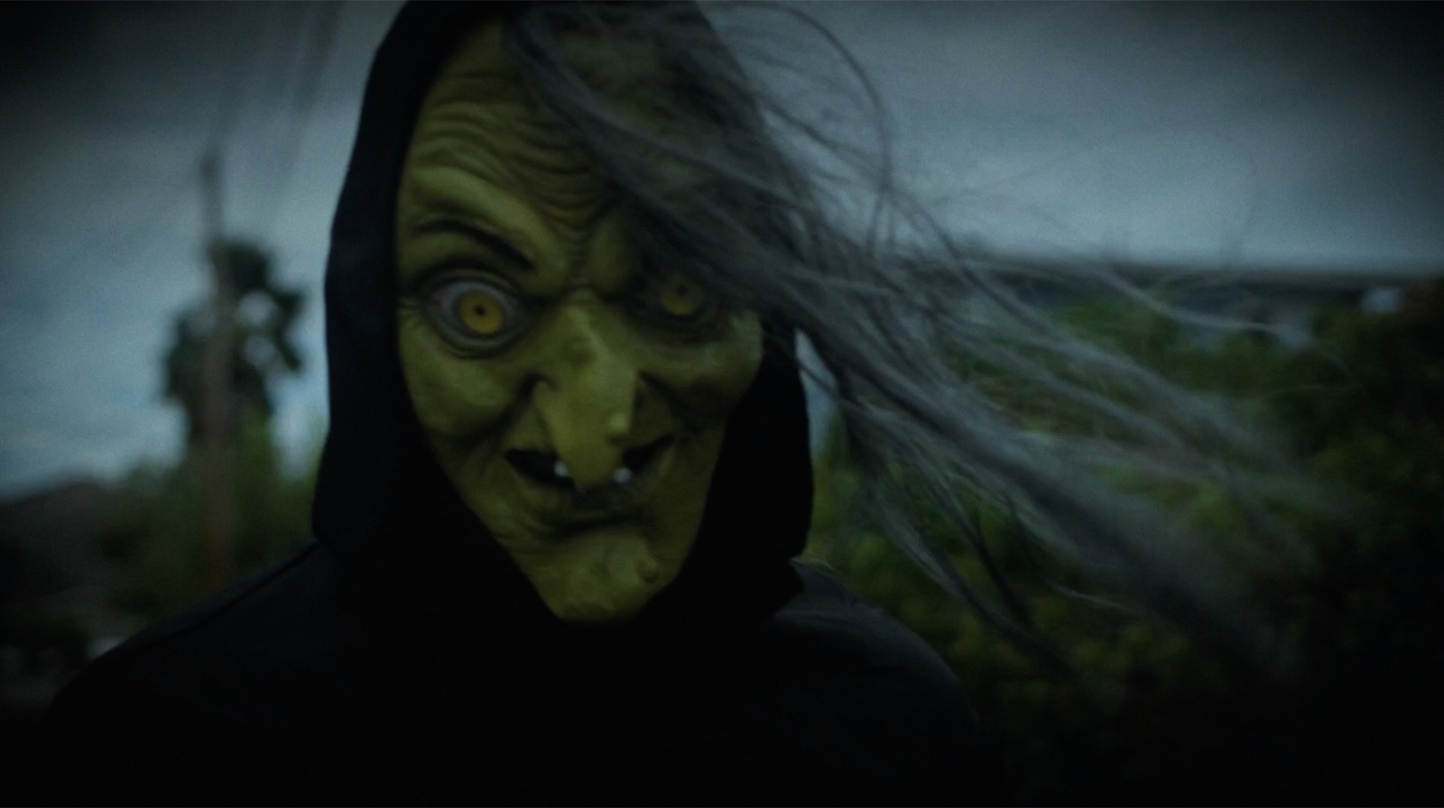 The witch stares into the camera, grey hair whipping in the wind under a storm-dark sky